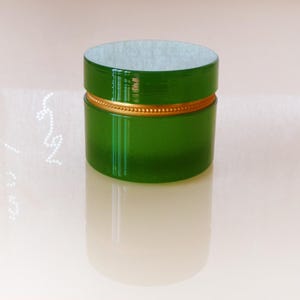 May include: A cylindrical, green container with a gold-colored band around the middle. The container has a lid with a white interior. The container is reflected in the surface below.