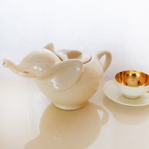 May include: A white ceramic teapot shaped like an elephant with a lid. The teapot is sitting on a white surface next to a white saucer with a gold-rimmed teacup.