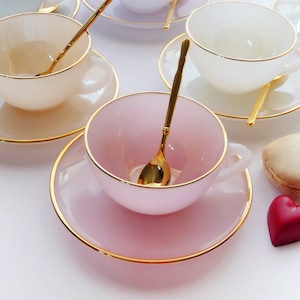 May include: A set of five pastel-coloured teacups and saucers with gold trim. Each cup and saucer has a matching gold-coloured spoon. The cups and saucers are arranged on a white surface.