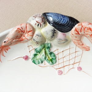 May include: A white ceramic serving dish with a raised design of two shrimp, a mussel, and various seashells. The dish is decorated with a green leafy design and a brown net pattern.
