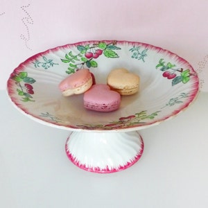 Può includere: Un porta torta in ceramica bianca con un design floreale rosa e verde. Il supporto ha un bordo ondulato e una base a piedistallo. Tre macarons a forma di cuore sono sul piatto. I macarons sono rosa e beige.