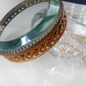 May include: An antique, gold-toned metal and glass bangle bracelet. The bracelet features a teal-colored glass band encased in a decorative, openwork metal frame. A clear glass dish is partially visible in the background.