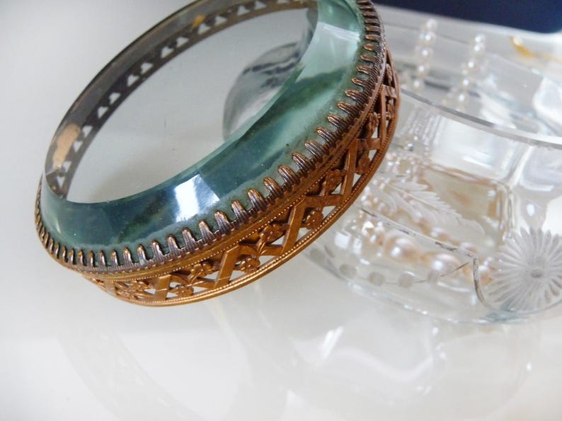 May include: An antique, gold-toned metal and glass bangle bracelet. The bracelet features a teal-colored glass band encased in a decorative, openwork metal frame. A clear glass dish is partially visible in the background.