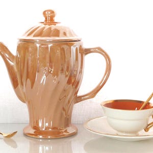 以下が含まれることがあります： 蓋、取っ手、注ぎ口が付いた、虹色のオレンジブラウンのセラミックティーポット。 金色の縁と取っ手が付いた白いティーカップがソーサーの上にあり、中に金色のスプーンが入っています。 2つ目の金色のスプーンが表面に置かれています。