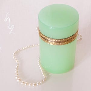 May include: A cylindrical, light green container with a gold-colored band around the middle. A delicate pearl necklace is draped around the container. The container has a smooth, glossy finish.