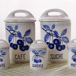 Könnte beinhalten: Ein Set aus fünf Keramik-Küchenbehältern mit Deckeln. Die weißen Behälter haben ein blaues Kirschzweig-Design und schwarze Textaufschriften: "CAFE", "SUCRE", "POIVRE", "GIROFLE" und "MUSCADE". Die Deckel haben einen kleinen Griff.