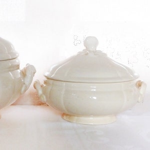 Peut inclure: Plat de service en céramique crème avec couvercle. Le plat a une forme arrondie avec une base à piédestal et deux poignées. Le couvercle est surmonté d'un bouton décoratif. Le plat est probablement vintage ou antique.