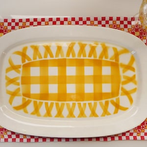 Könnte beinhalten: Eine weiße ovale Servierplatte mit einem gelben und weißen karierten Muster in der Mitte. Das karierte Muster ist von einem gelben Rand aus ineinander verschlungenen Bändern umgeben.