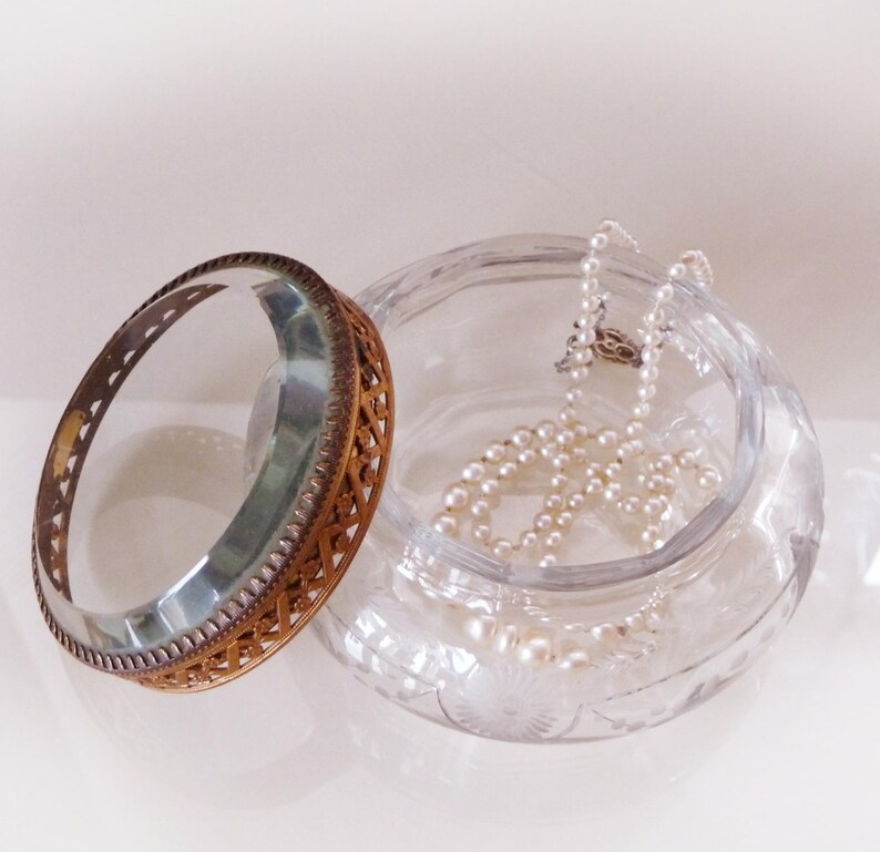 May include: A clear glass jewelry box with a decorative lid and a pearl necklace inside. The lid is made of metal with an intricate openwork design. The box has a round shape and is sitting on a white surface.
