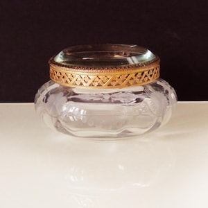 May include: A clear glass jar with a decorative gold-colored band around the rim. The jar has a clear glass lid and is sitting on a white surface. The jar has etched floral designs.