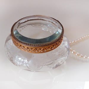May include: A clear glass inkwell with a decorative etched floral design. The inkwell has a gold-colored metal rim and a magnifying glass. A strand of pearls is draped nearby.