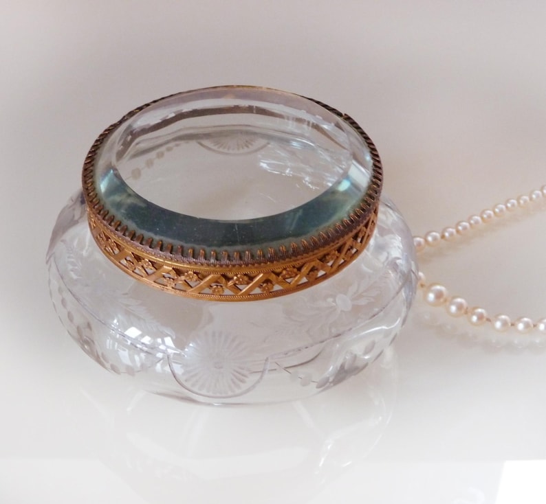 May include: A clear glass inkwell with a decorative etched floral design. The inkwell has a gold-colored metal rim and a magnifying glass. A strand of pearls is draped nearby.