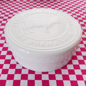 Può includere: Piatto per burro in ceramica bianca con un design circolare in rilievo che presenta una silhouette di mucca e il testo "Verrerie de Tomme d'Anjou".