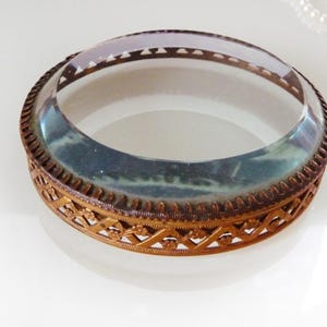May include: An antique-style, round glass and metal decorative piece. The clear glass top is surrounded by a bronze-colored metal frame with an intricate, openwork design. The piece sits on a white surface.