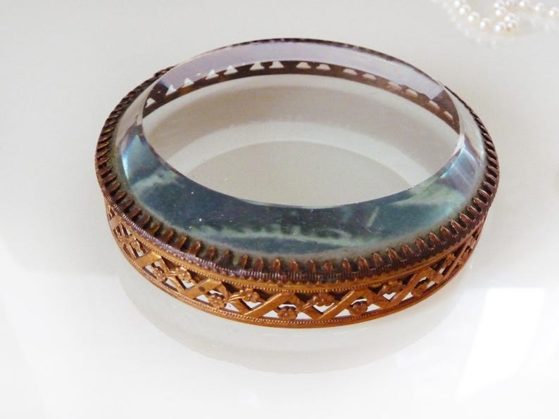 May include: An antique-style, round glass and metal decorative piece. The clear glass top is surrounded by a bronze-colored metal frame with an intricate, openwork design. The piece sits on a white surface.