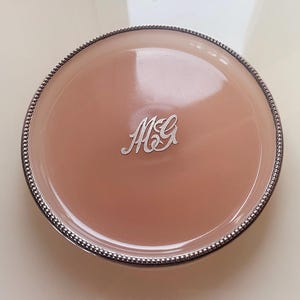 May include: A round, peach-colored compact with a silver beaded edge and a silver monogram. The compact has a shiny, reflective surface. The monogram features the letters "M" and "G" in a stylized script.