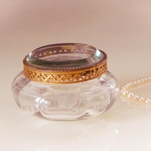 May include: A clear glass jar with a gold filigree rim and a clear glass lid. A strand of pearls rests beside the jar. The jar has a floral design etched into the glass.