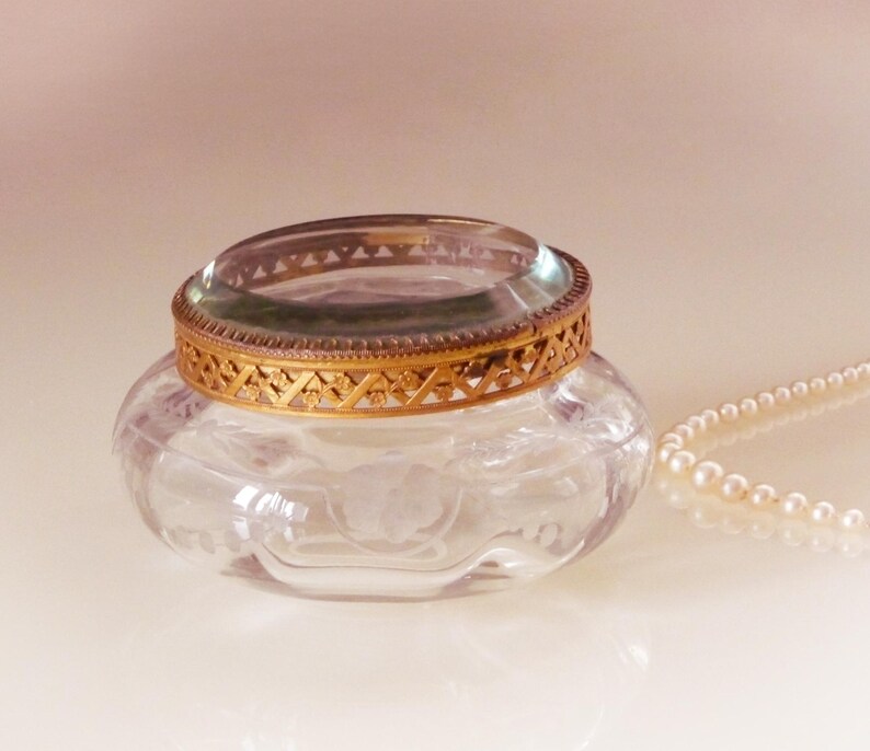 May include: A clear glass jar with a gold filigree rim and a clear glass lid. A strand of pearls rests beside the jar. The jar has a floral design etched into the glass.