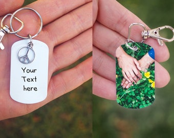 Könnte beinhalten: Zwei silberne Schlüsselanhänger mit unterschiedlichen Designs. Der erste Schlüsselanhänger hat einen Friedenszeichen-Anhänger und den Text "Your Text here". Der zweite Schlüsselanhänger hat ein Foto von zwei Händen, die sich auf einem Bett aus grünem Gras halten.