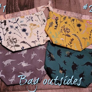 Pode incluir: Quatro sacos com cordão com diferentes padrões. Dois sacos têm desenhos florais em creme e amarelo. Os outros dois sacos apresentam silhuetas de dinossauros em cinza e verde-azulado. O texto "Bag outsides" está escrito na parte inferior.