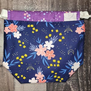 May include: A blue drawstring bag with a floral print featuring pink and white flowers, yellow leaves, and gold stars. The bag has a purple lining with a white star pattern.