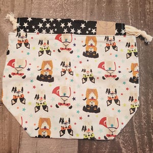 May include: A drawstring pouch featuring a white base with a repeating pattern of cartoon dogs wearing superhero capes and masks, interspersed with small colourful stars. The top section is black with a white star pattern and cream drawstrings.
