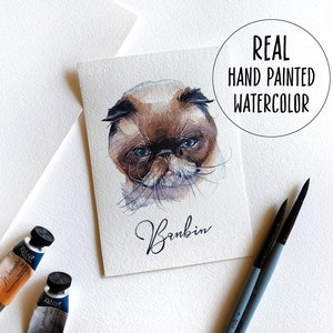 K&ouml;nnte beinhalten: Ein Aquarellbild eines Katers mit blauen Augen. Der Kater ist braun und wei&szlig; und hat ein flauschiges Gesicht. Das Bild ist mit "Bambin" signiert und der Text "REAL HAND PAINTED WATERCOLOR" befindet sich in einem Kreis oben im Bild.