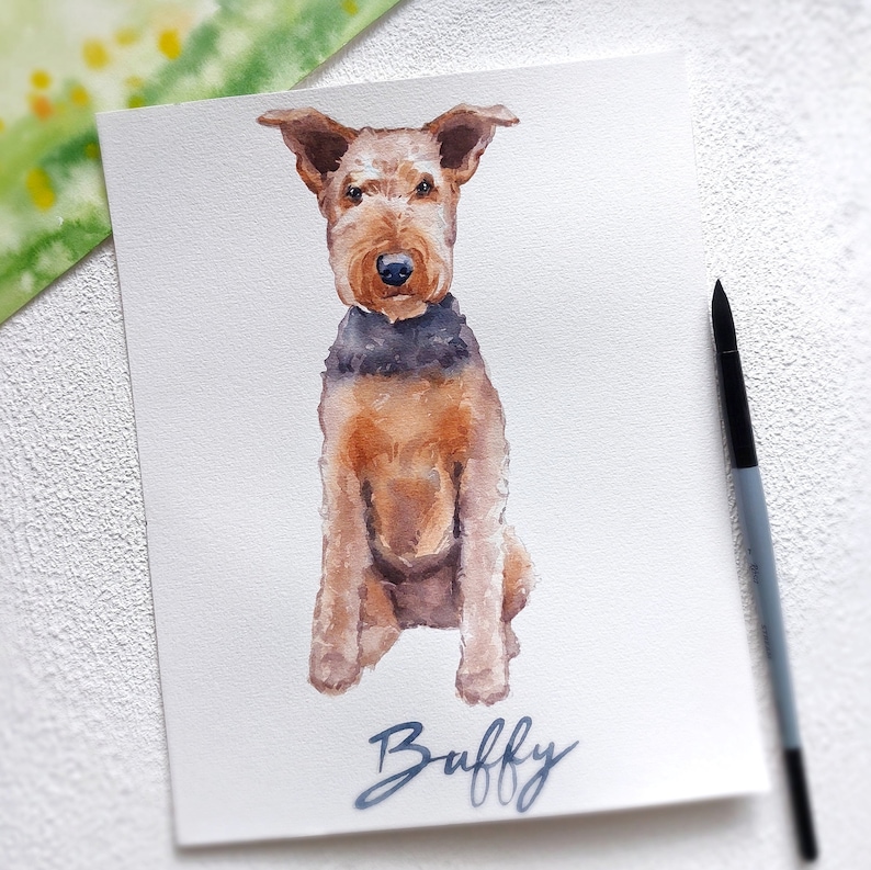 K&ouml;nnte beinhalten: Aquarellmalerei eines braunen und schwarzen Airedale Terrier Hundes, der sitzt und den Betrachter ansieht. Der Name des Hundes ist "Buffy" und steht in blauer Kursivschrift unter dem Hund.
