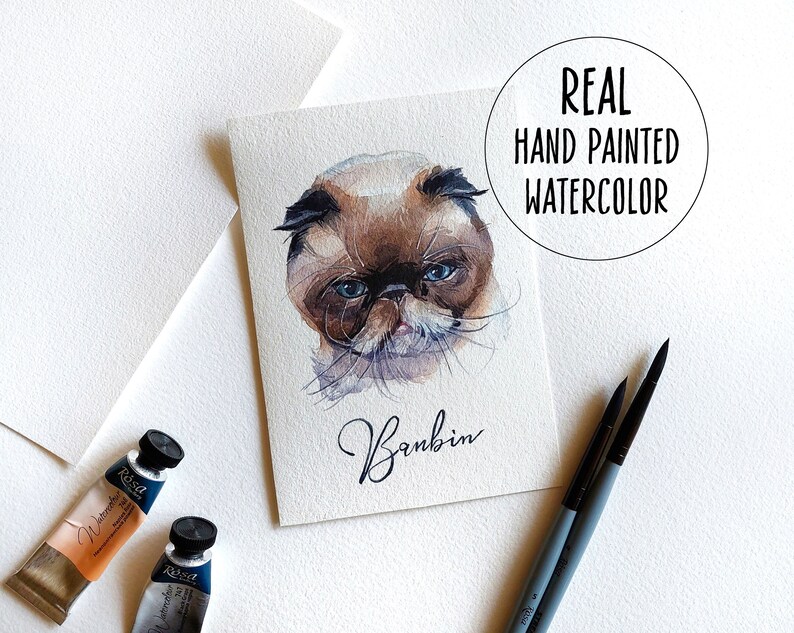 May include: A watercolour painting of a cat with blue eyes. The cat is brown and white and has a fluffy face. The painting is signed "Bambin" and the text "REAL HAND PAINTED WATERCOLOUR" is in a circle at the top of the image.