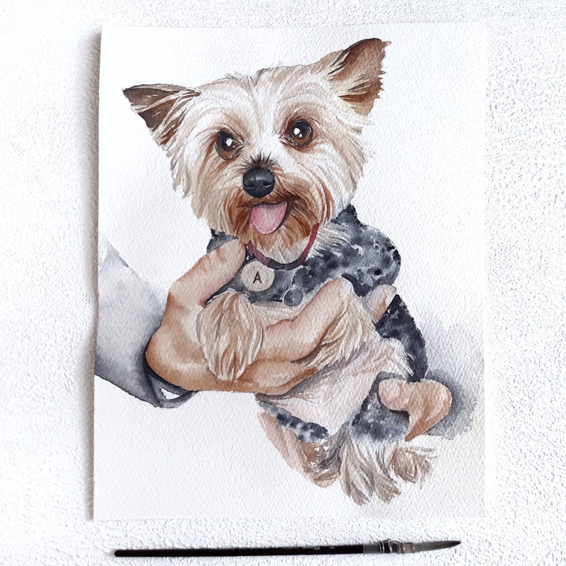 May include: Watercolour painting of a small brown and black Yorkshire Terrier dog being held in a hand. The dog is wearing a black collar with a silver tag that says "A".