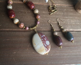 May include: A set of gemstone jewelry featuring a long necklace with a large oval pendant and matching earrings. The necklace is made with brown, beige, and pink stones and gold accents. The earrings are made with brown, beige, and green stones and gold accents.