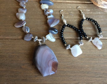 May include: A necklace and earrings set featuring a large, gray agate pendant with a silver clasp. The necklace is made of smaller agate beads and black beads. The earrings are silver hoops with black beads and a small agate pendant.