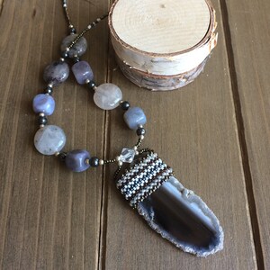 Peut inclure: Un collier avec un grand pendentif en agate gris, entouré d'une bordure de perles. Le collier est fait de perles grises, blanches et brunes, et d'une perle de cristal.