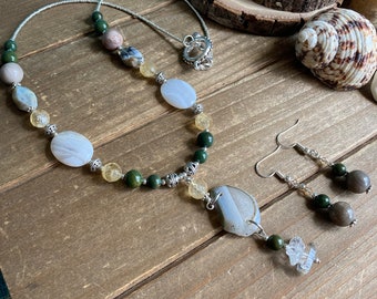 May include: A silver necklace with a large, light gray agate pendant. The necklace is made with various beads, including green, yellow, and white. The necklace is paired with matching earrings with green and brown beads.