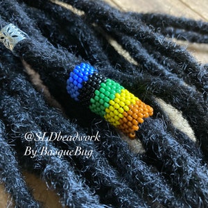 May include: A colorful beaded dreadlock bead with blue, black, green, yellow, and brown beads. The bead is on a set of black dreadlocks. The text "@SJDbeadwork By BasqueBug" is visible in the image.