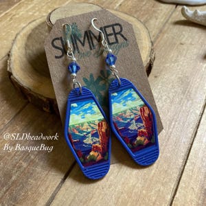 May include: A pair of blue and silver earrings featuring a vintage motel key tag design with a picture of the Grand Canyon. The earrings have blue crystal accents.