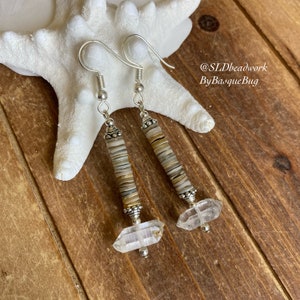 May include: A pair of silver earrings with a long, dangling design. The earrings feature a stack of small, round, beige and brown beads, topped with a clear quartz crystal. The earrings are hanging from a silver hook.