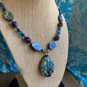 May include: A necklace featuring a large abalone shell pendant with a wire-wrapped tree of life design. The necklace is made with abalone shell beads, amethyst beads, and copper wire.