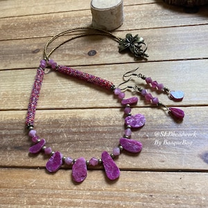 May include: A pink and gold beaded necklace with a pink stone pendant and matching earrings. The necklace features a beaded section with a pink and gold colour scheme. The pendant is a large, irregularly shaped pink stone. The earrings are small and feature a pink stone with a gold wire loop. The necklace and earrings are on a wooden surface. @SL.Dbeadwork By BasqueBug