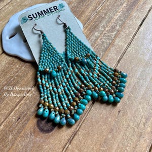 Puede incluir: Pendientes de aro de cuentas turquesa y doradas con un diseño de flecos. Los pendientes están en una tarjeta que dice "Summer Love Designs" y "@SLDbeadwork By Basque Bug".