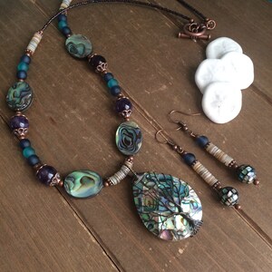 May include: A copper wire wrapped abalone shell pendant necklace and earrings set. The necklace features abalone shell beads, amethyst beads, copper beads, and blue glass beads. The earrings feature abalone shell beads, copper beads, and blue glass beads.