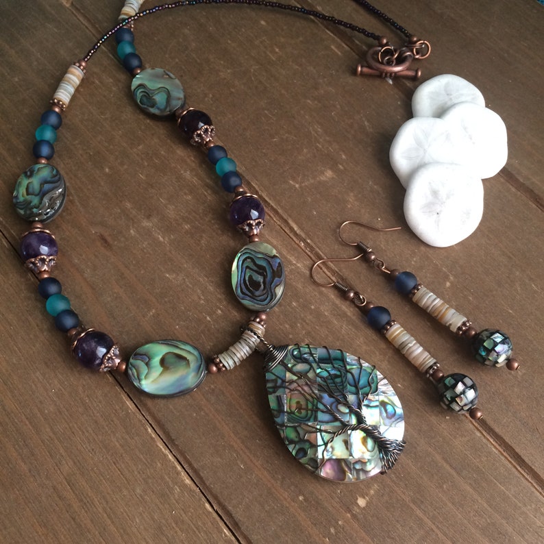 May include: A copper wire wrapped abalone shell pendant necklace and earrings set. The necklace features abalone shell beads, amethyst beads, copper beads, and blue glass beads. The earrings feature abalone shell beads, copper beads, and blue glass beads.