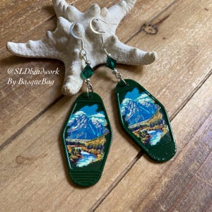 May include: Green and silver earrings featuring a vintage motel key tag with a mountain scene. The key tag is green with a white border and a picture of a mountain range with a river in the foreground. The earrings are hanging from silver hooks with green crystal beads.