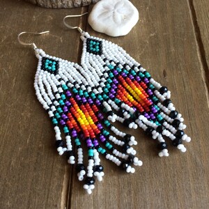 May include: A pair of long, beaded earrings with a colorful geometric pattern. The earrings are made with white, black, turquoise, purple, orange, yellow, and red beads. The earrings have silver hooks.