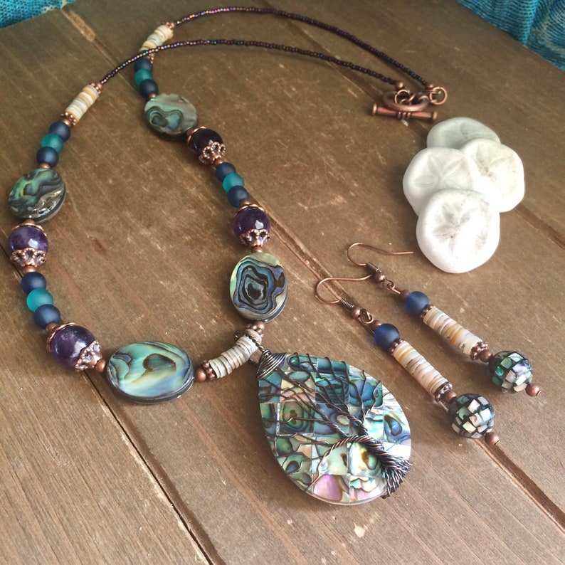 May include: A necklace and earrings set featuring a teardrop-shaped pendant with a wire-wrapped tree design. The pendant and beads are made of abalone shell, with copper accents and blue and white glass beads.