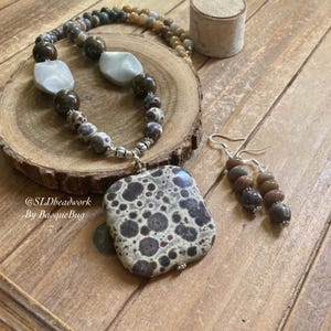 May include: A brown and white speckled stone pendant necklace with matching earrings. The necklace has a silver chain and the pendant is a square shape. The earrings are silver with a small brown and white speckled stone on each earring.