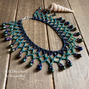 May include: A beaded necklace with a graduated color scheme of dark blue, teal, and green. The necklace has a geometric pattern and features iridescent teardrop-shaped beads. The necklace is on a wooden surface with a seashell.