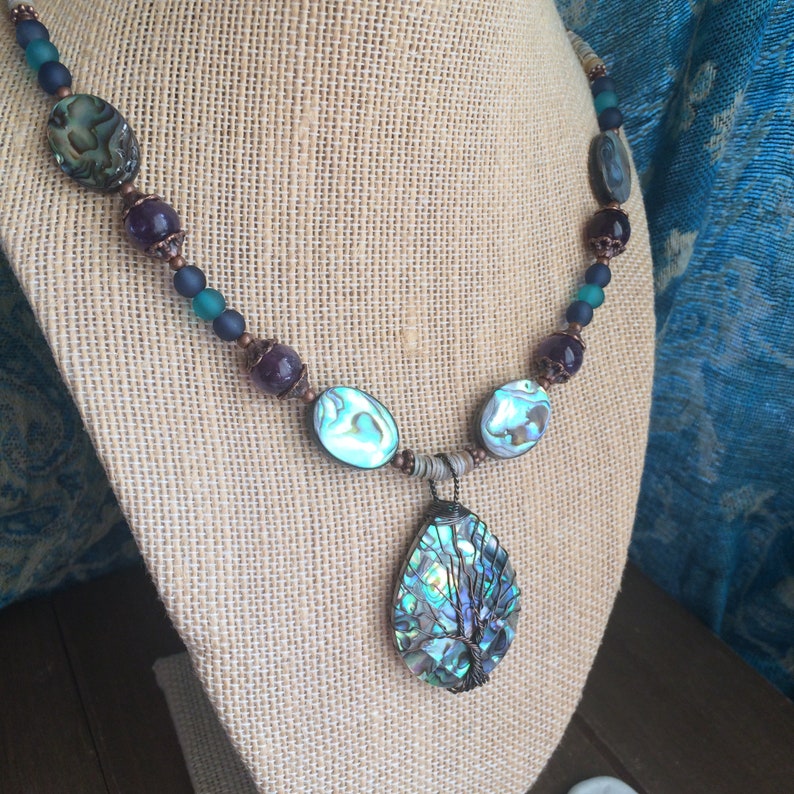 May include: A necklace with a large abalone shell pendant featuring a wire-wrapped tree design. The necklace is made with copper wire, abalone shell beads, amethyst beads, and blue glass beads.