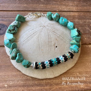 May include: Turquoise gemstone bracelet with a black, white, and gold beaded center section. The bracelet has a silver clasp and is shown on a light brown wooden surface.
