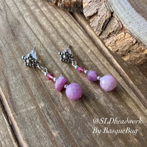 Puede incluir: Un par de pendientes colgantes con cuentas rosas y transparentes y detalles plateados. Los pendientes están sobre una superficie de madera. El texto "@SLDbeadwork By BasqueBug" es visible en la esquina inferior derecha de la imagen.